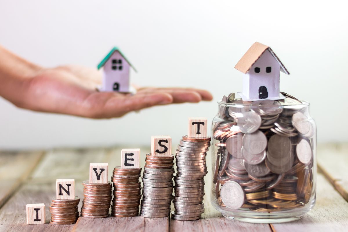 The photo of a jar filled with coins and with a mini house, coins stocks with wooden cubes and letters, and a man's hand holding a mini house refers to investing in assets that appreciate over time and provide financial freedom for you and your family.