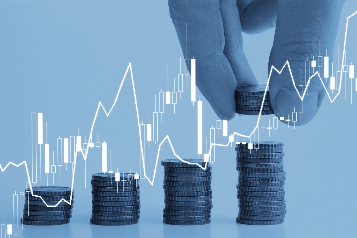The picture shows coin stocks, visual lines and graphs, and a man's hand putting coins on the last coins tower referring to diversified investments increasing financial stability and reducing the chances of losing wealth.