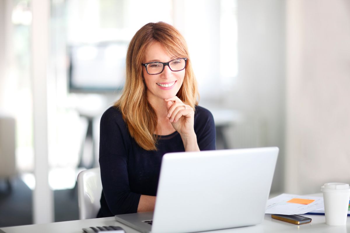 The image shows a coffee, a phone, papers, and a girl using a laptop while smiling referring to digital product arbitrage is a low-risk way to make money from anywhere.