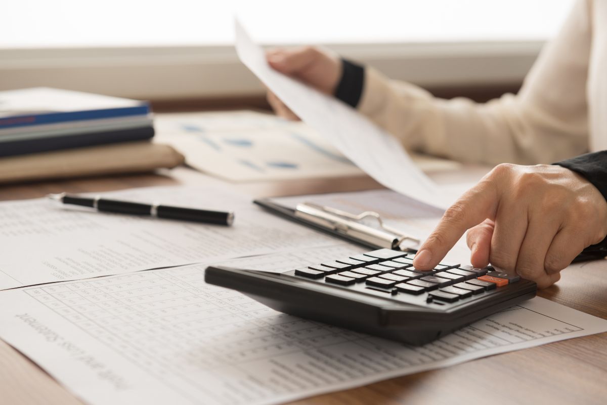 The image shows notebooks, papers on the table, a pen, and a girl's hand calculating while holding a paper which refers to passing down financial knowledge to future generations and helps them make smart financial decisions.