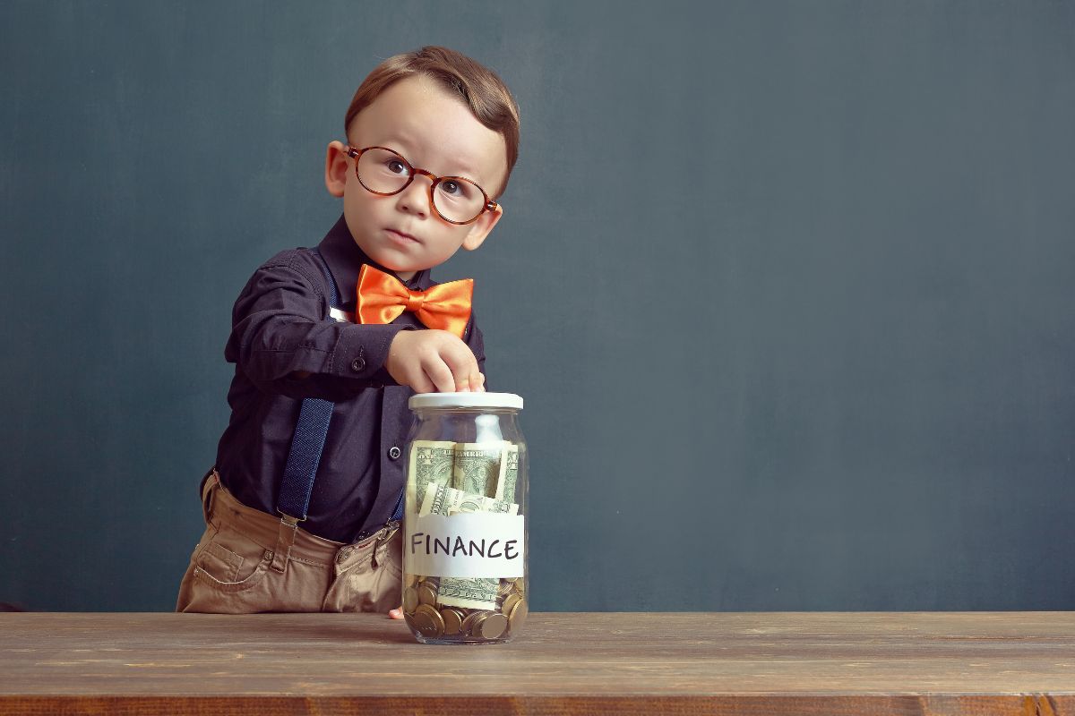 The picture of a young boy putting money inside a jar filled with dollar bills, and coins, and with a note indicating to teach children about money management early helps ensure your wealth grows over generations.