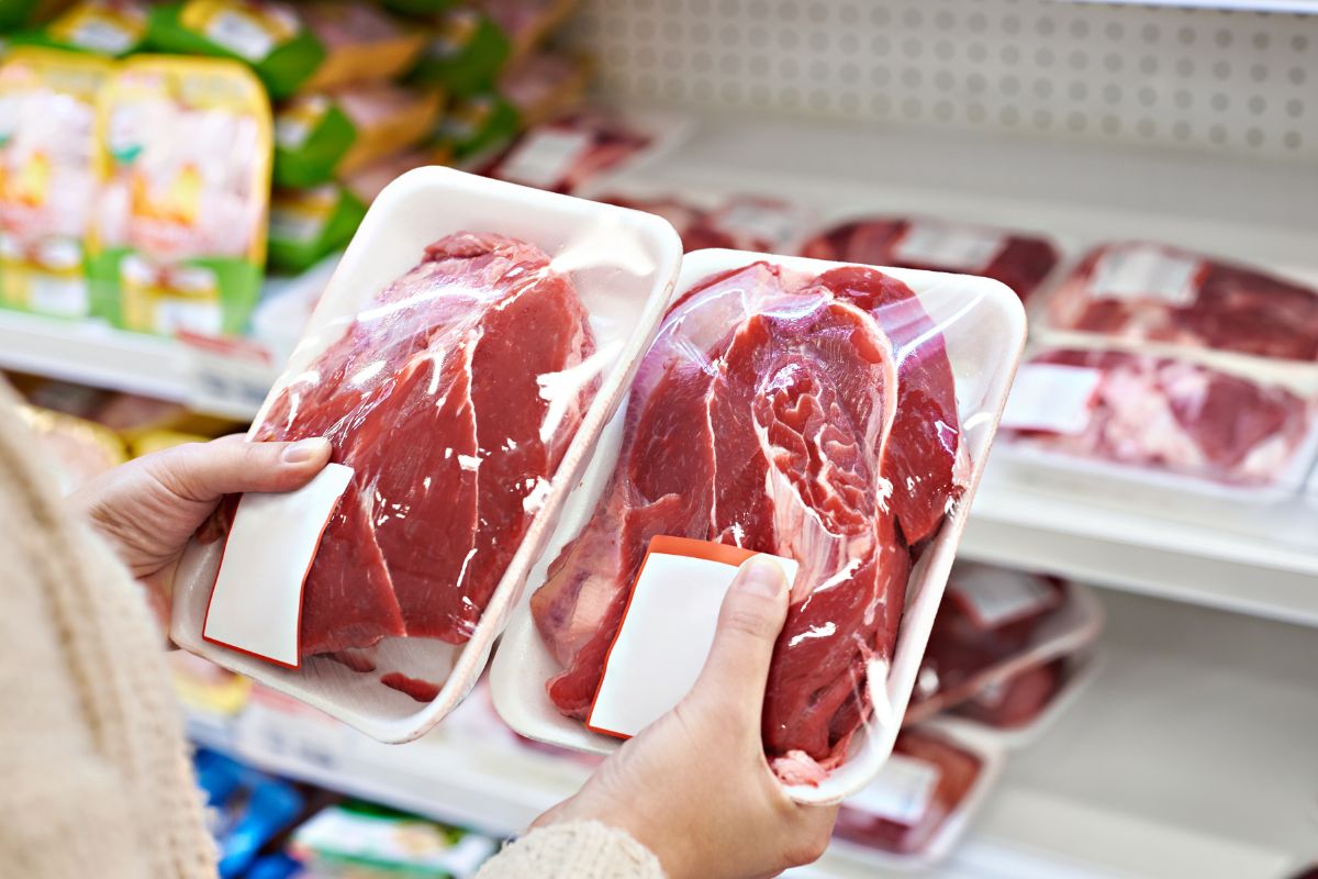 The picture shows a girl's hand picking up two packaged meats at the store indicating that purchasing whole cuts of meat is a long-term way to save money on groceries.