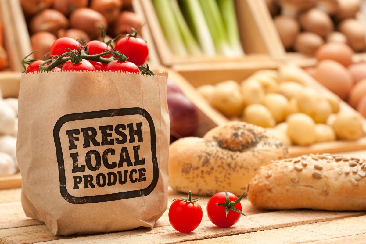 The picture shows vegetables in a wooden box, bread, and a paper bag with tomatoes inside referring to joining local farm Co-Ops programs that help avoid waste and keep costs down long-term.