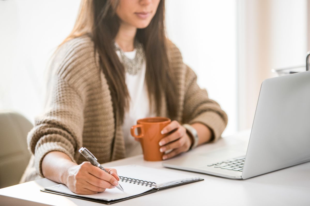 The image tells about a laptop and a girl writing in the notebook while having a coffee referring to front-loading your high-value tasks early in the day means getting more done before distractions kick in.