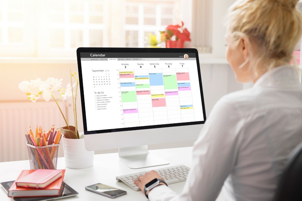 The photo tells about pens, a notebook, a phone, a tablet device, and a girl using a computer with a screen display referring to implementing reverse scheduling to get more than in less time while maintaining balance.