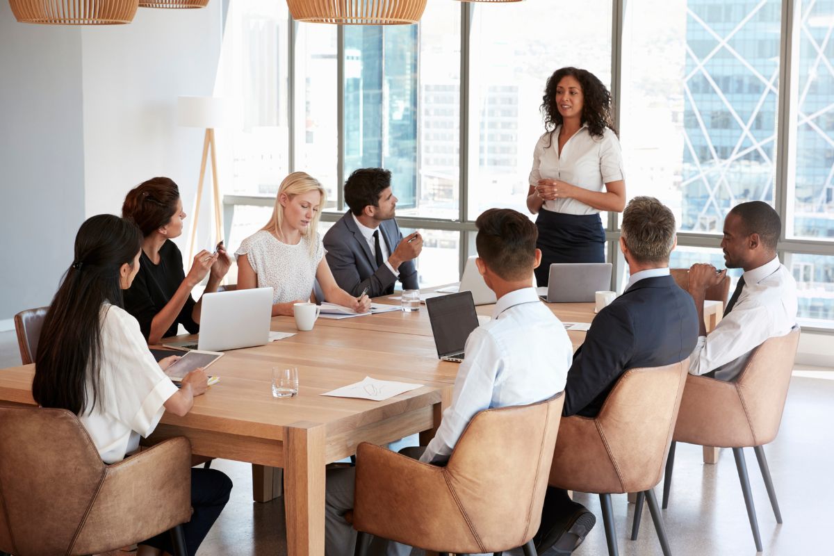 The photo tells about a group of employees sitting while listening to the speaker with their laptops and papers to take notes indicating that adopting a no-meeting mornings policy helps people get more done and earn more in the long run.