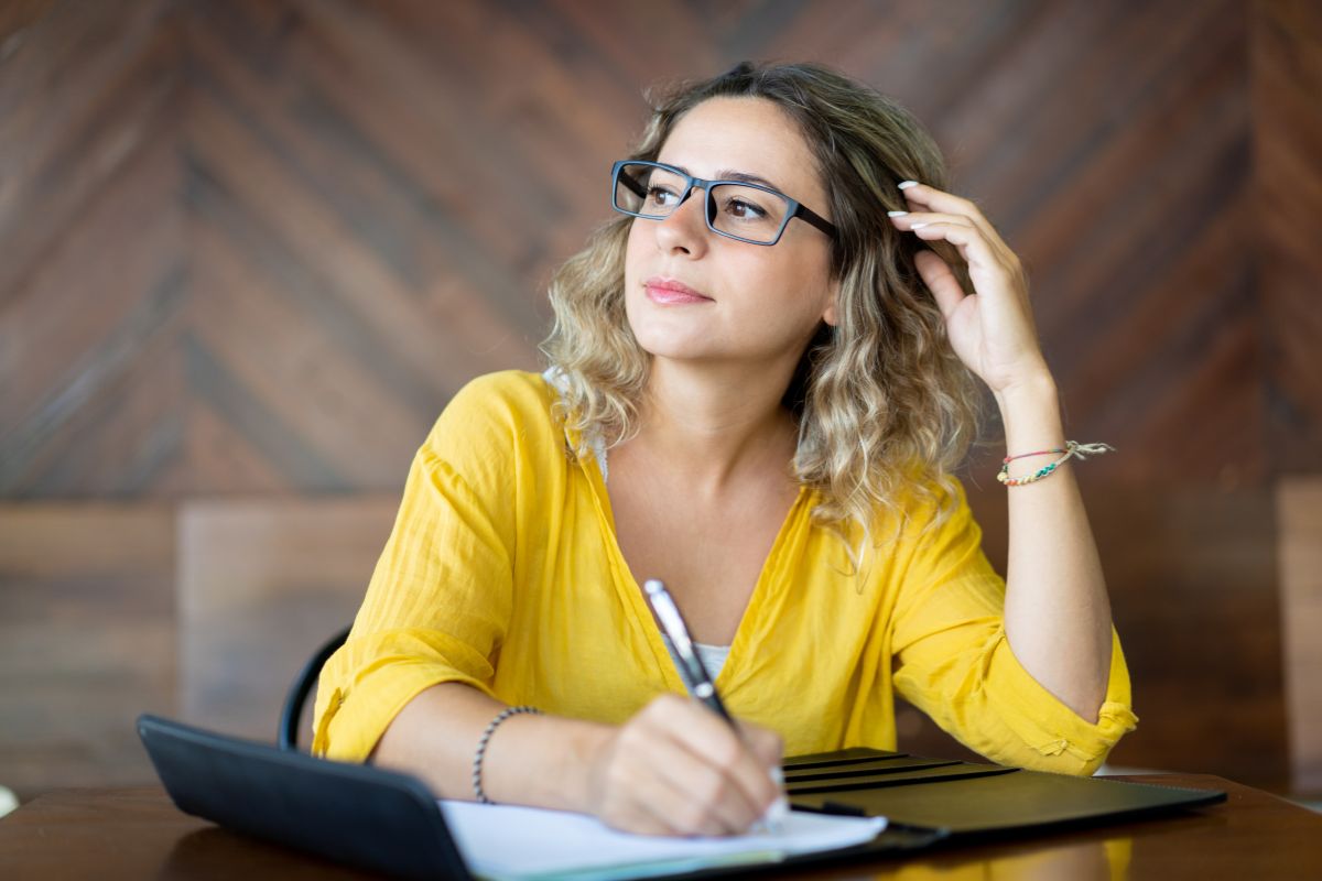 The photo shows a girl writing in the notebook while looking outside preferring to use an accountability mirror to keep financial goals top of mind and help measure daily progress.
