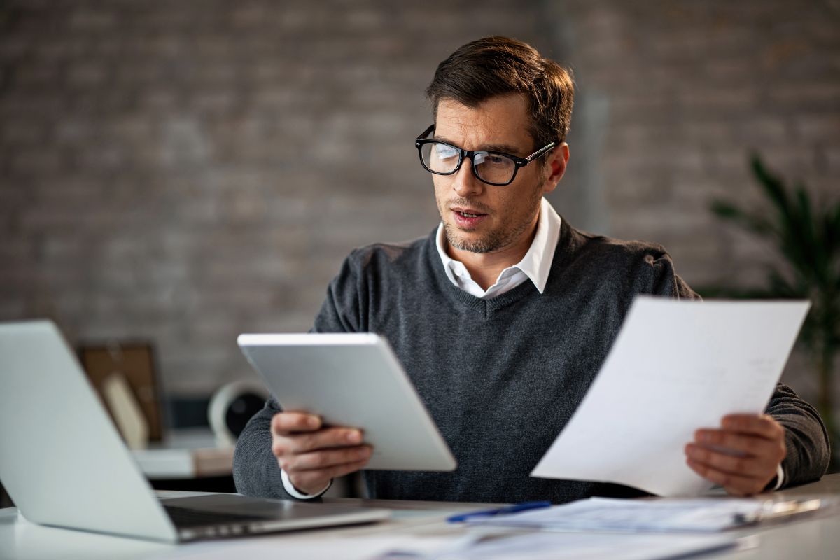 The photo shows a laptop, papers on the table, and a man holding a tablet device and a piece of paper which refers to outsourcing aggressively value tasks leading to better productivity and stronger financial results.