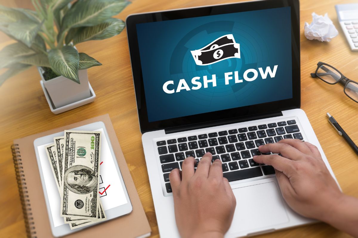 The picture tells about a plant, a notebook, a tablet device, dollar bills, an eyeglass, a calculator, a pen, and a man's hand using a laptop with a screen display indicating that setting up cash-flow checkpoints keeps focus on revenue-generating activities and helps avoid financial surprises.