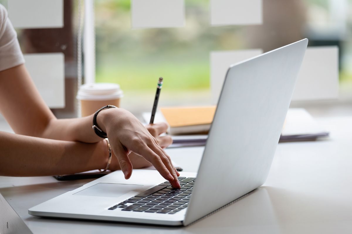 The picture tells about a coffee, papers, and a girl's hand using a laptop while holding a pen referring to online reputation management for individuals who can bring in steady clients who value their online image.