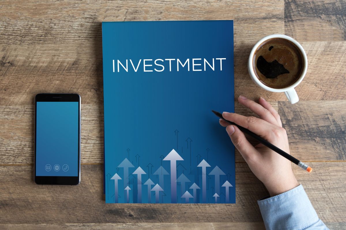 The image shows a coffee, a phone, a book with a note and arrows, and a man's hand holding a pen referring to an investment in knowledge that pays the best interest and has a return that never stops growing.