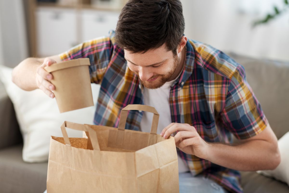 The photo tells about a man opening a paper bag with packed food indicating that takeout and food delivery can cost much, coking at home helps control portion sizes and save money. 