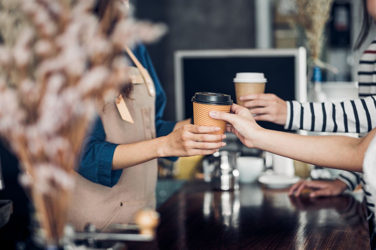 The picture tells about a counter's hand handing the coffee to the customers which refers to coffee shop visits adding up fast, making coffee at home can cut costs without giving up the habit.