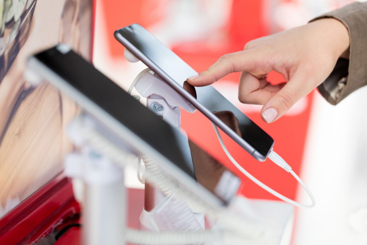 The photo shows a girl's hand checking on new phones at the store referring to unnecessary tech upgrades, holding onto gadgets longer saves money in the long run.