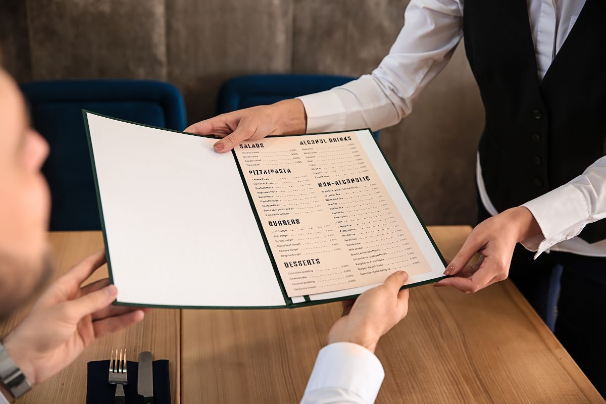 The picture shows a waiter's hand handing the menu to the customer which refers to over-ordering at restaurants makes bills climb fast, drinking water instead of soda helps save money. 