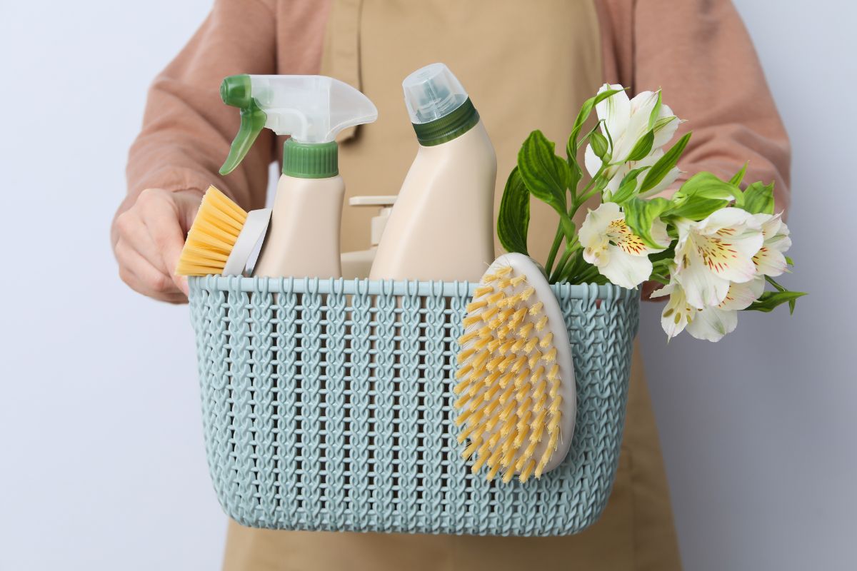 The image tells about a girl's hand carrying a basket with flowers and cleaning tools indicating that fancy cleaning products cost more than necessary, and choosing multipurpose cleaners over specialty ones also saves money.