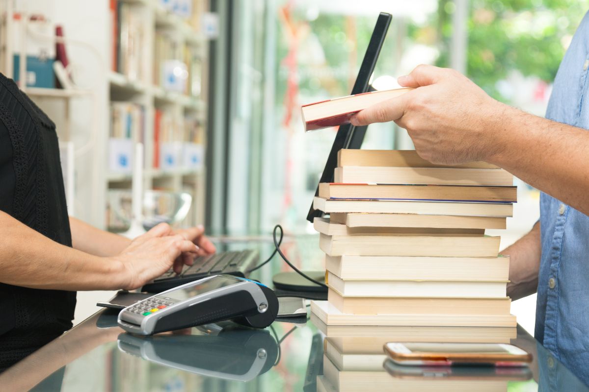 The image tells about books on the table, a phone, and a man's hand showing a book on the counter who type in the computer with a paying machine which means buying books gets expensive fast, borrowing helps cut down on clutter and spending.