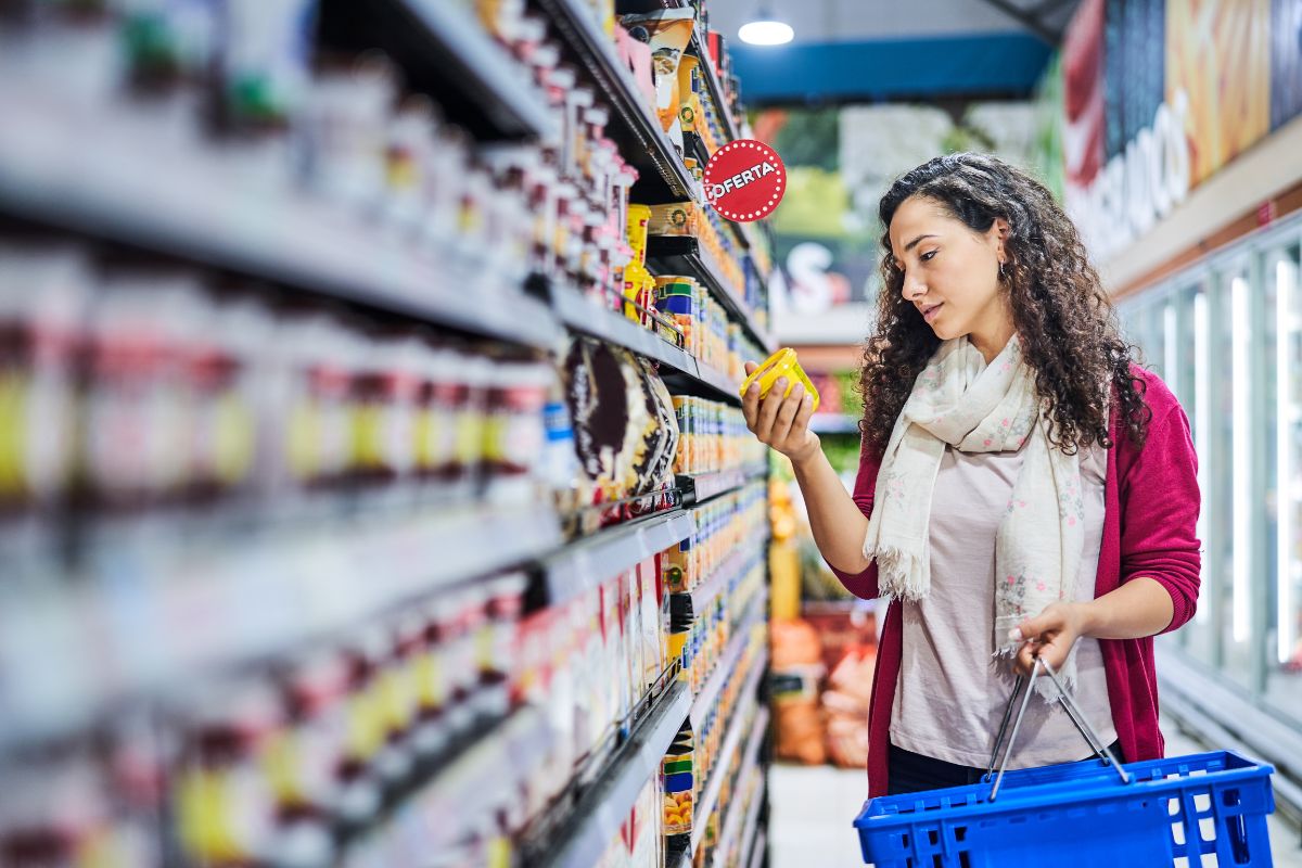 The picture shows a product display and a girl picking one product while holding a basket which refers to buying full-price items may feel urgent, but waiting for sales can save a lot of money.