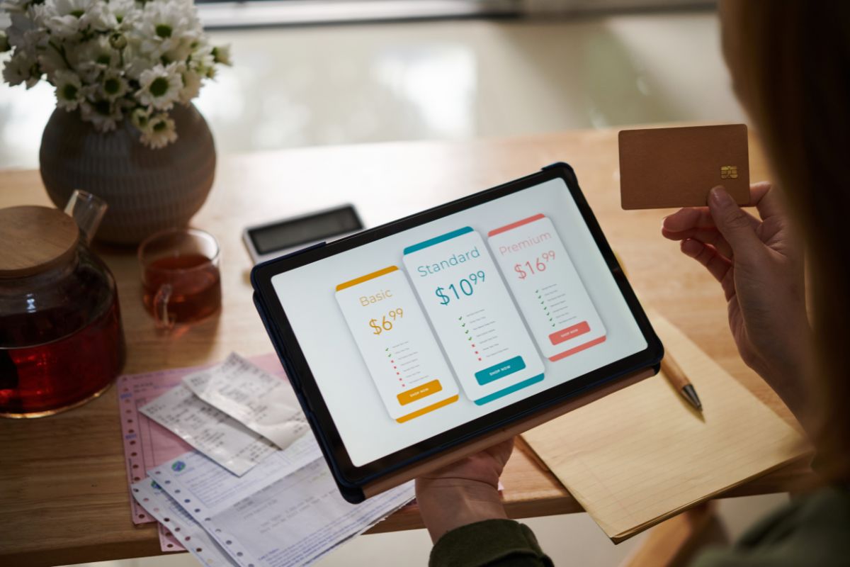 The image tells about a flower vase, tea, papers on the table, papers with a pen, and a girl's hand holding a tablet device and a credit card referring to multiple streaming services, canceling services that offer redundant content and sticking to one or two can save money.