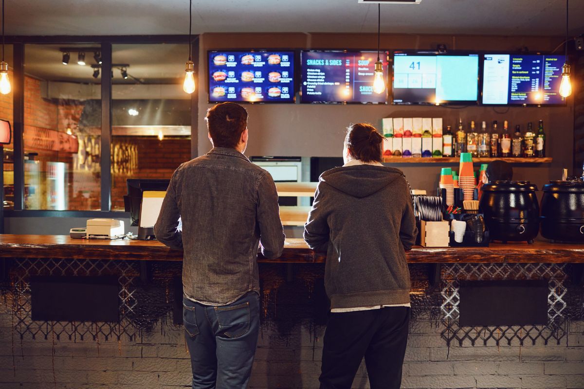 The photo shows customers ordering food at the counter referring to spending more on convenience than necessity leads to wasted money, cooking at home can help keep finances on track.