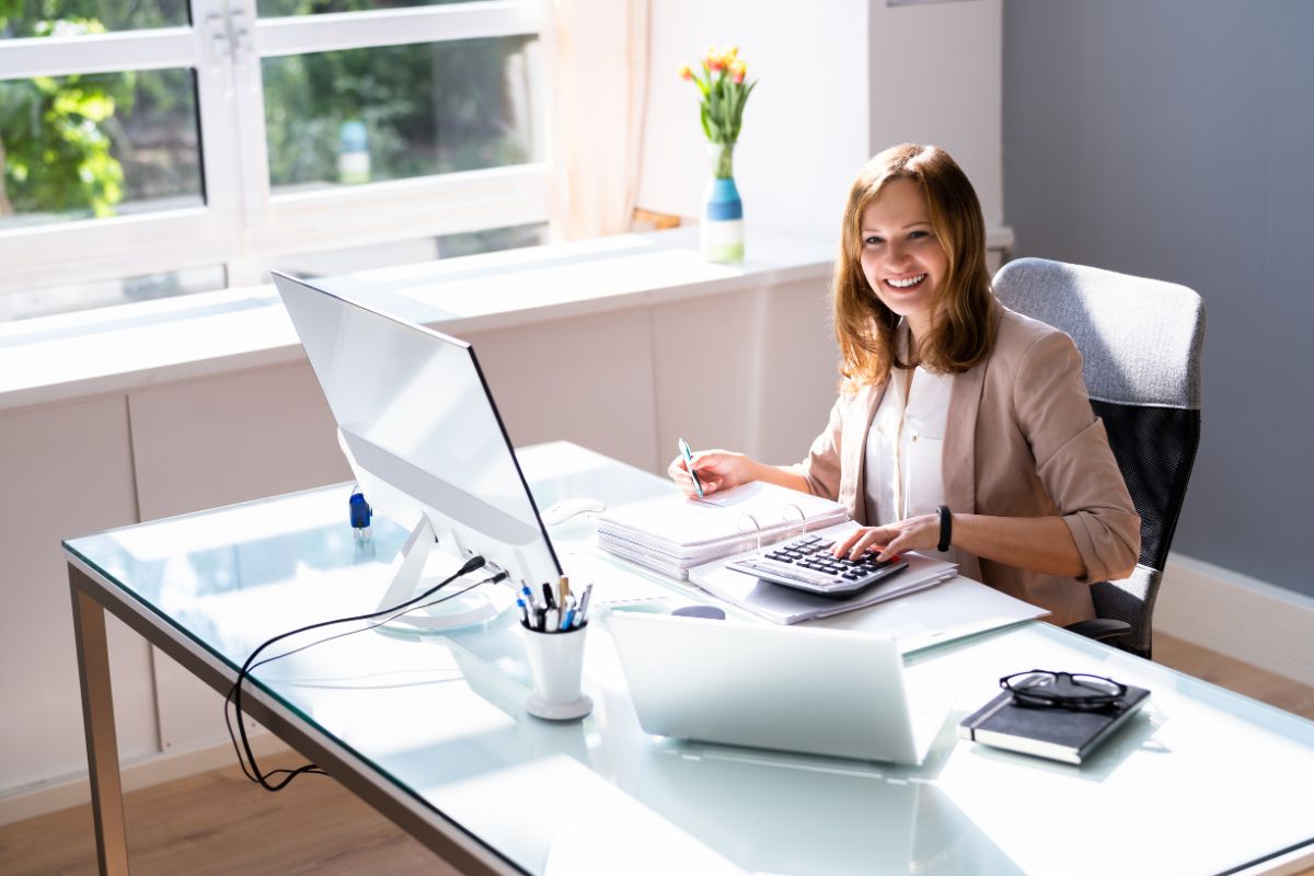 The picture of a notebook, an eyeglass, pens, a laptop, a computer, and a girl writing on the papers while calculating which refers to remote bookkeeping and accounting services is steady work-from-home with good income potential.