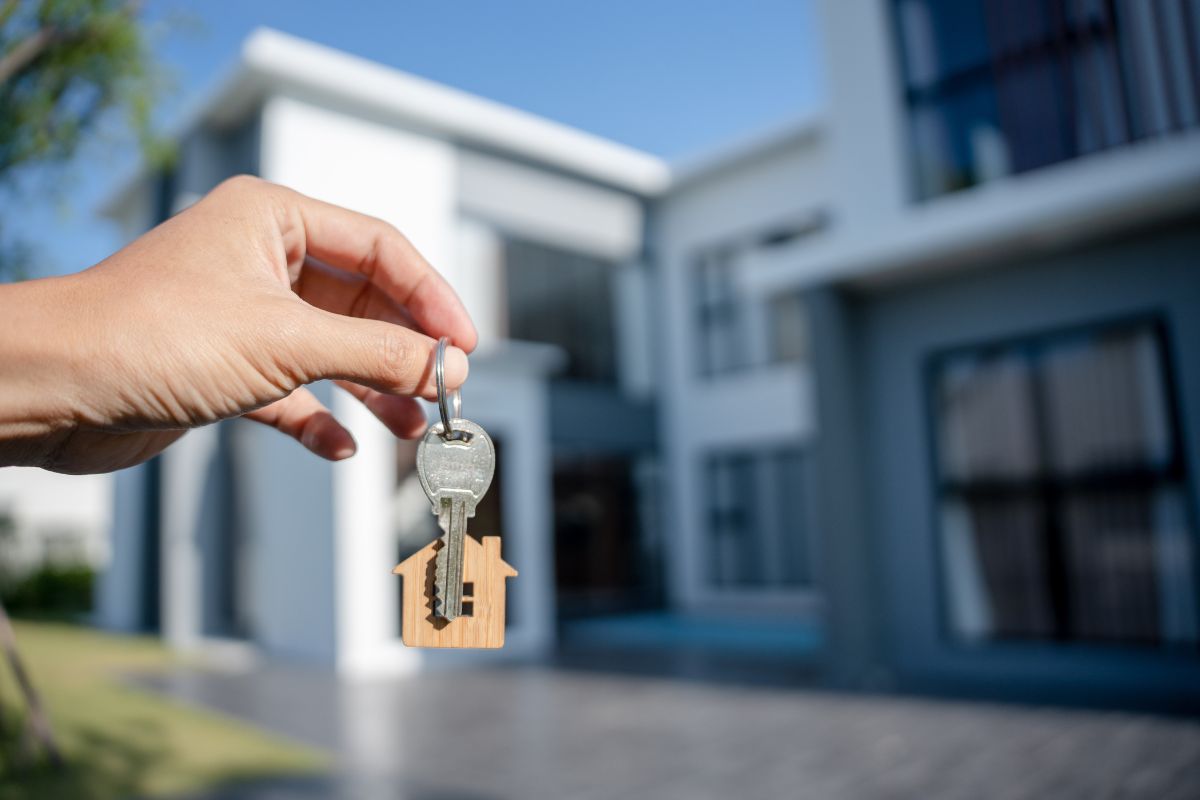 The image shows a man's hand holding a house key and a house behind which refers to buying an expensive home too early leading to stress, renting or buying within budget allows for more financial flexibility.