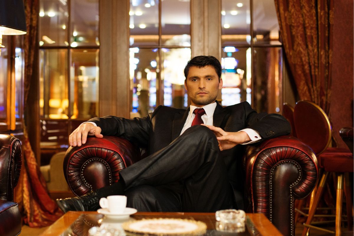 The picture of a man in a suit sitting on the couch while having a coffee referring to rich people's plans and thinking beyond the next paycheck can change your entire financial path.
