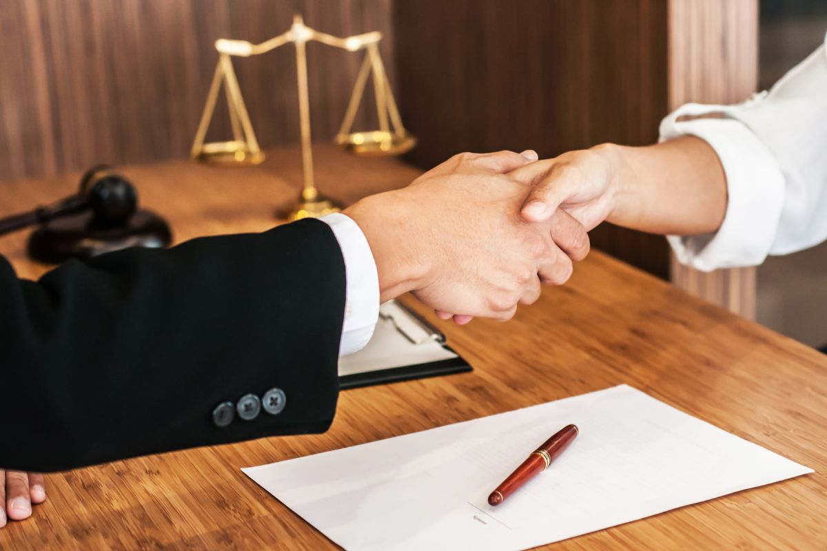 The photo tells about a justice display, a clipboard, a pen and paper, and two people shaking hands together which means corporate lawyers help businesses with contracts that pay well.