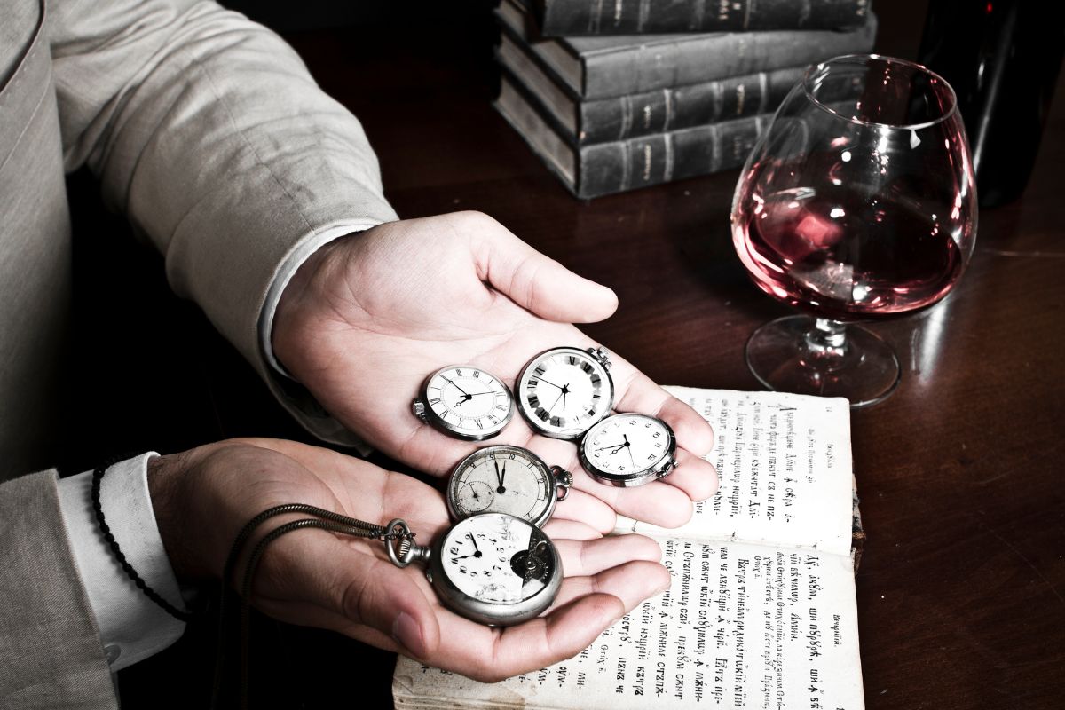 The photo shows books, a drink, and a man's hand holding vintage clocks which refers to rare antique dealers and selling them for profit.