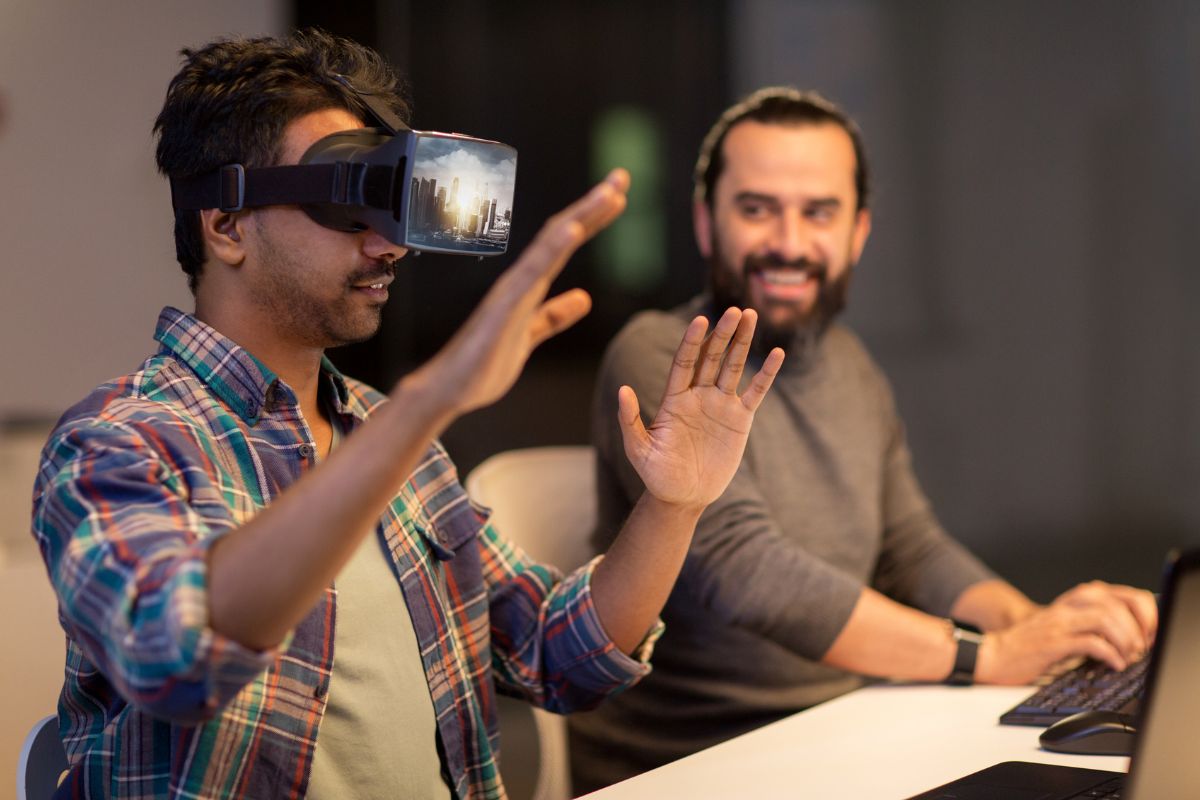 The photo shows a man typing on the computer and a man testing the VR which refers to virtual reality developers building apps and interactive experiences with a high-income career.