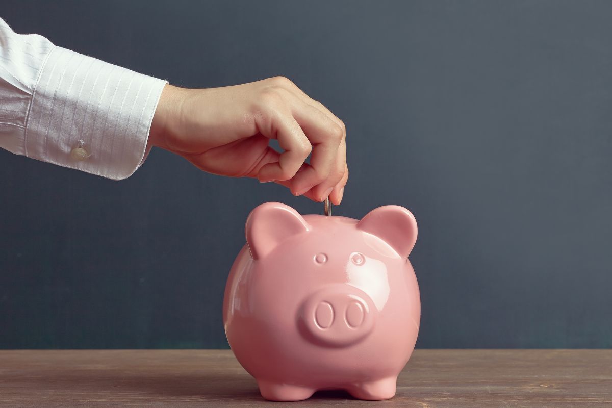 The photo shows a man's hand dropping a coin in a piggy bank which refers to yourself first method to help build savings fast and make sure financial goals come first.