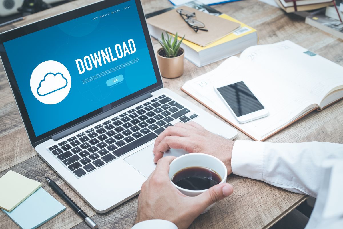 The image shows a plant, notebooks, a phone, a pen, and a man's hand using a laptop while having a coffee which refers to downloading your budgeting template printables that keep you in control of your money.