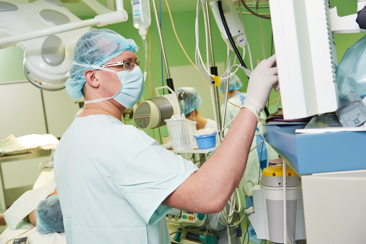 The picture shows a man in white scrubs with a mask and hairnet adjusting the machine inside an operating room together with his colleagues referring to an anesthesiologist who plays a key role in surgery that is a stable and high-paying medical career.