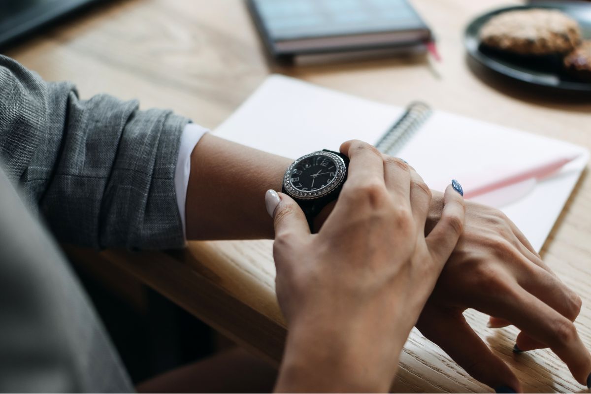 The photo of a meal in a plate, notebooks, a pen, and a girl's hand checking her watch which means time management keeps you from feeling overwhelmed later in life.