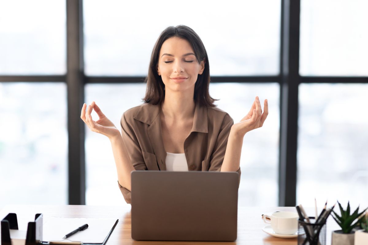 The picture shows a plant, a coffee, pens, papers, and a laptop, and a girl smiling while relaxing referring to stress management and mindfulness help prevent burnout means fewer health issues later.
