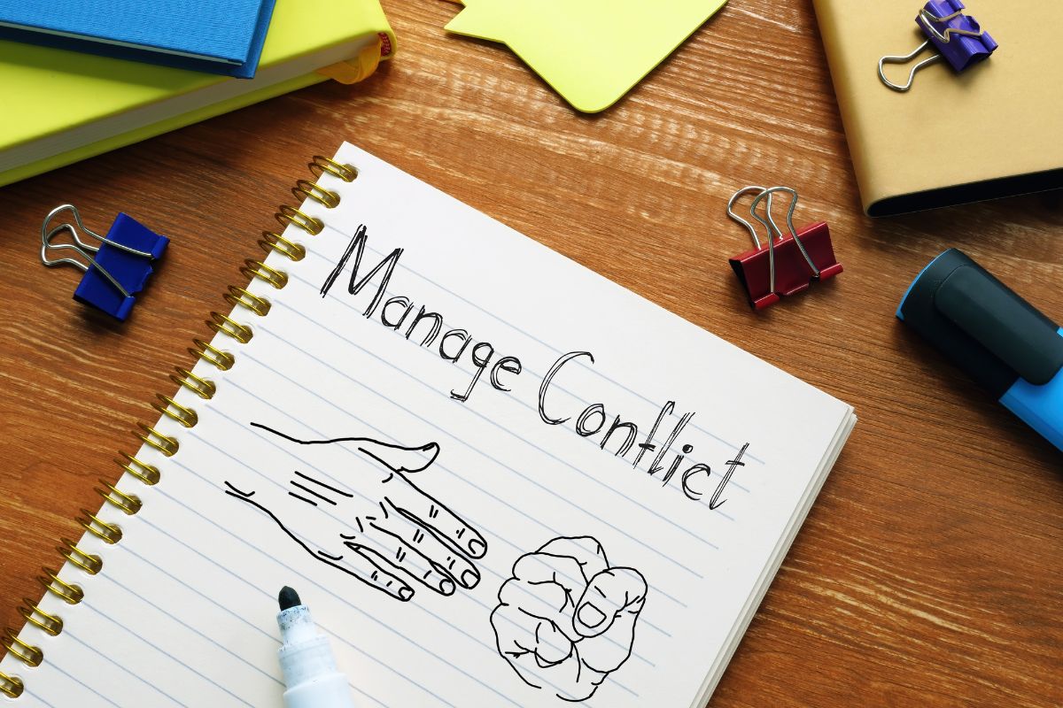 The picture shows notebooks, clips, a marker, and a notebook with notes indicating that conflict resolution builds trust and improves teamwork.
