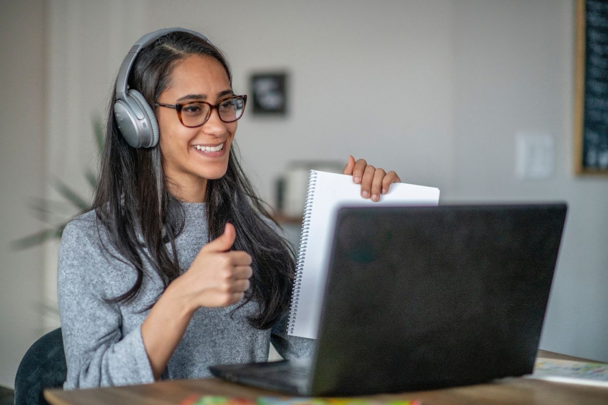 The photo shows a girl holding a notebook while making an ok sign and looking at the camera which refers to work as a side hustle for teachers like online teachings can bring in extra income without taking over your life. 