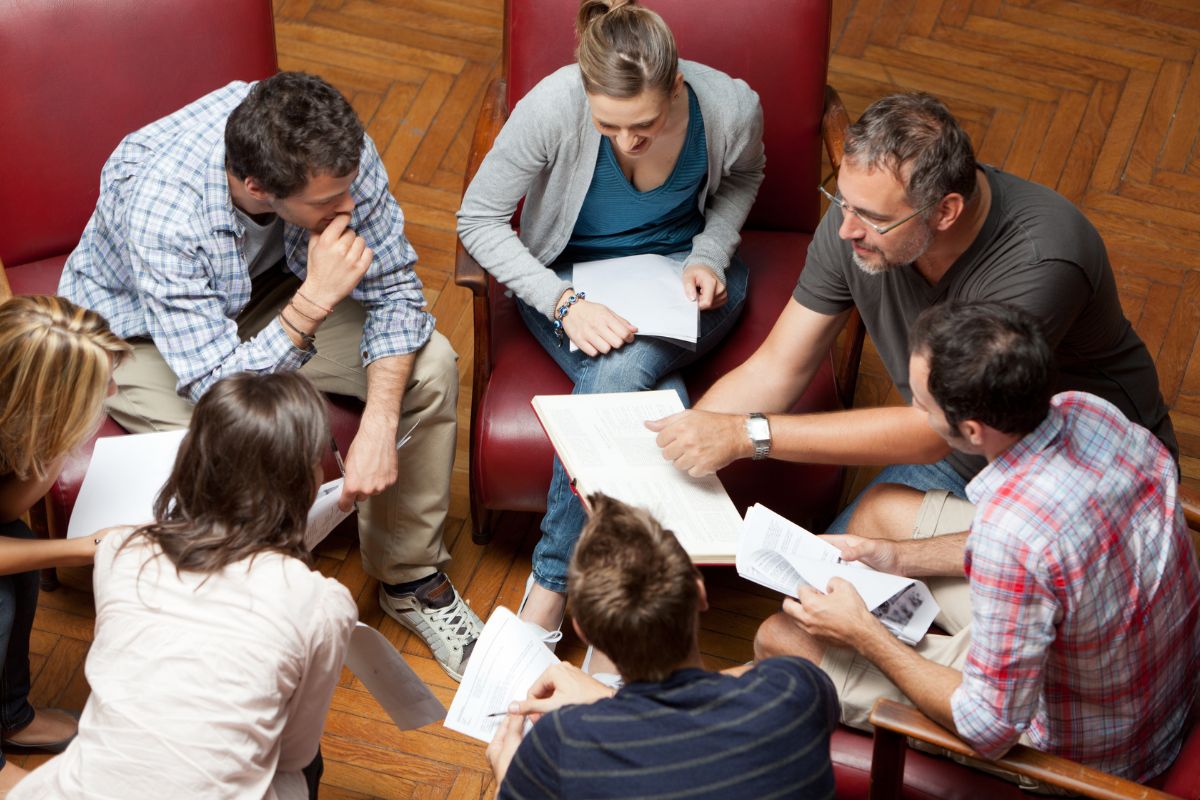 The photo shows a group of people holding papers while discussing it together indicating that staying local and doing things with friends makes it easy to balance work and fun.
