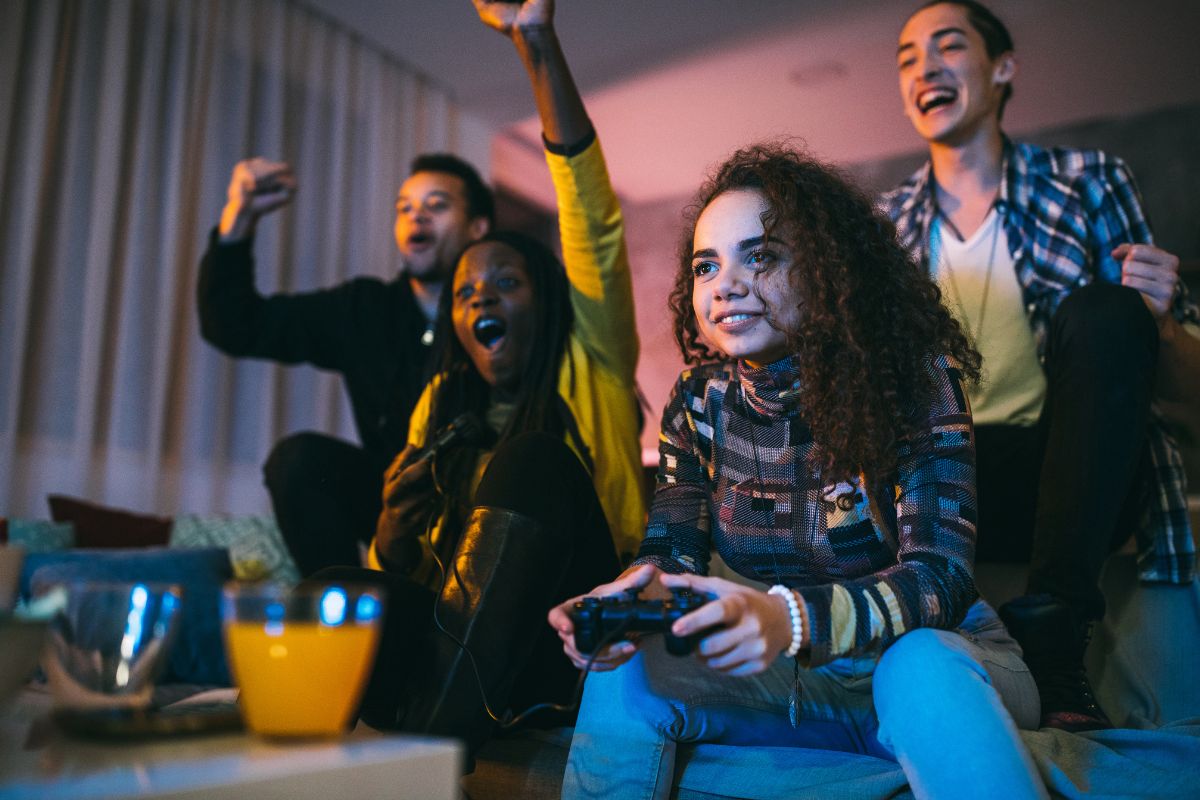 The image shows an orange drink and a group of young people playing video games which refers to turning fun money into challenge money making frugal living feel rewarding instead of restrictive.
