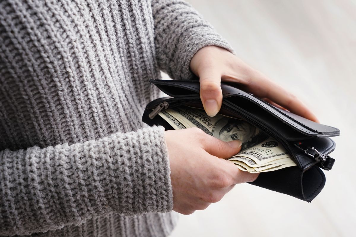 The picture shows a girl's hand putting back the dollar bills inside her wallet referring to keeping a no-spend day helps reset spending habits and makes it clear what's really needed.