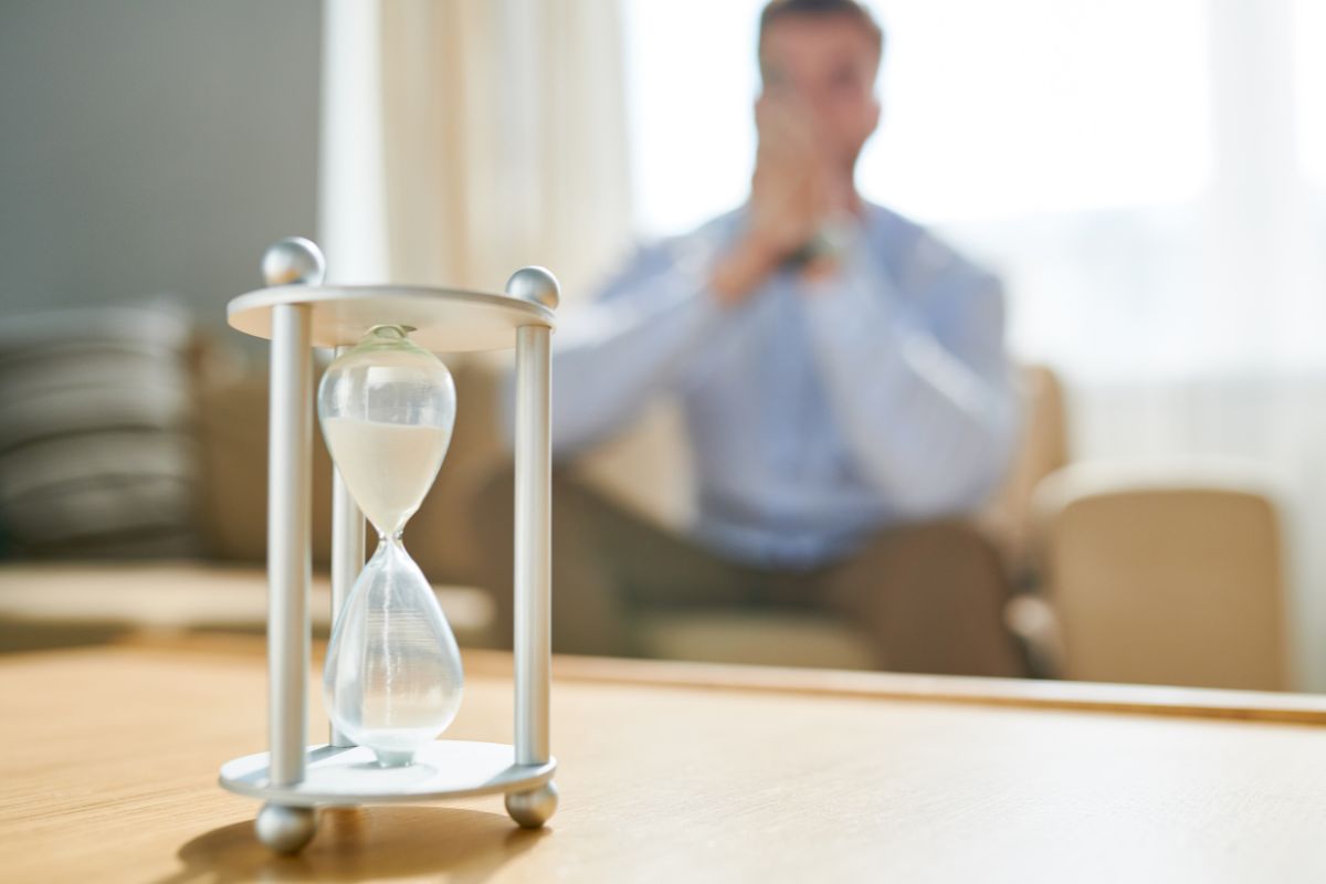 The image of a man sitting on the couch while looking at the glass hour referring to the wait-and-earn trick makes it easier to say no to impulse buys.