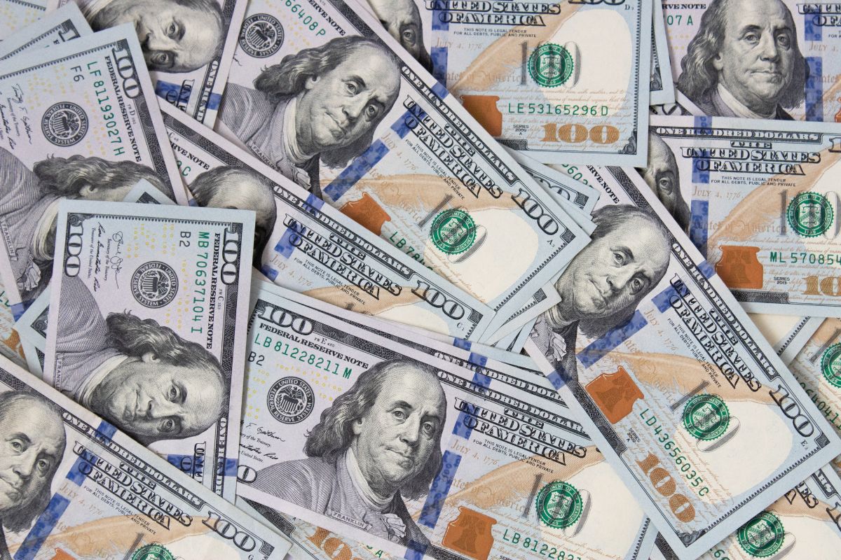 The picture shows scattered dollar bills referring to building up your first $100k saved and reaching this goal means having a strong safety net and more freedom in financial choices.