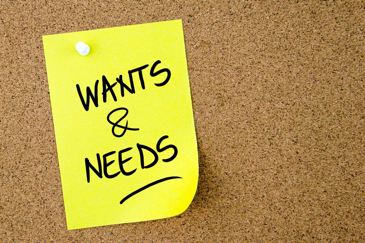 The picture of a piece of paper with notes pinned on the wall indicates the difference between wants and needs keeps more money in savings and helps reach financial goals sooner.
