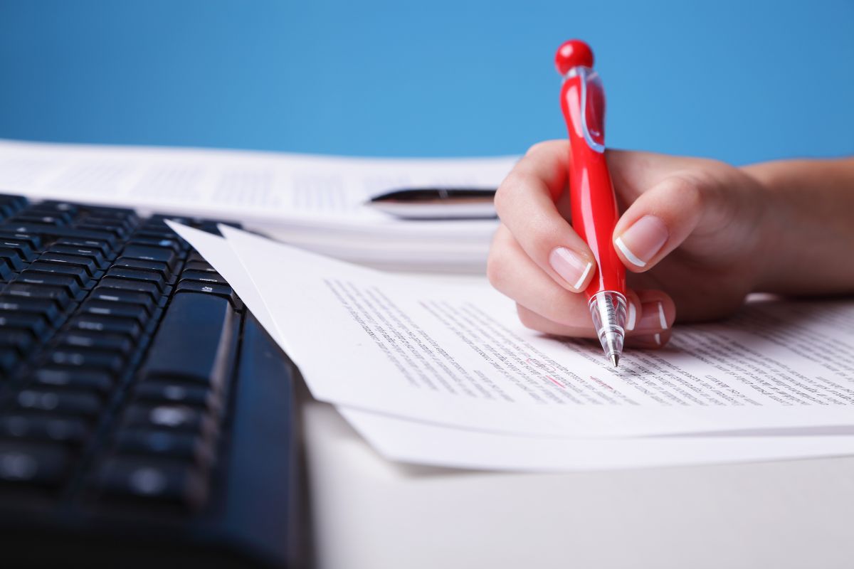 The picture shows a keyboard and a girl's hand holding a pen while checking on the papers with notes referring to proofreading as the right side hustle that could be a smart way to make money.