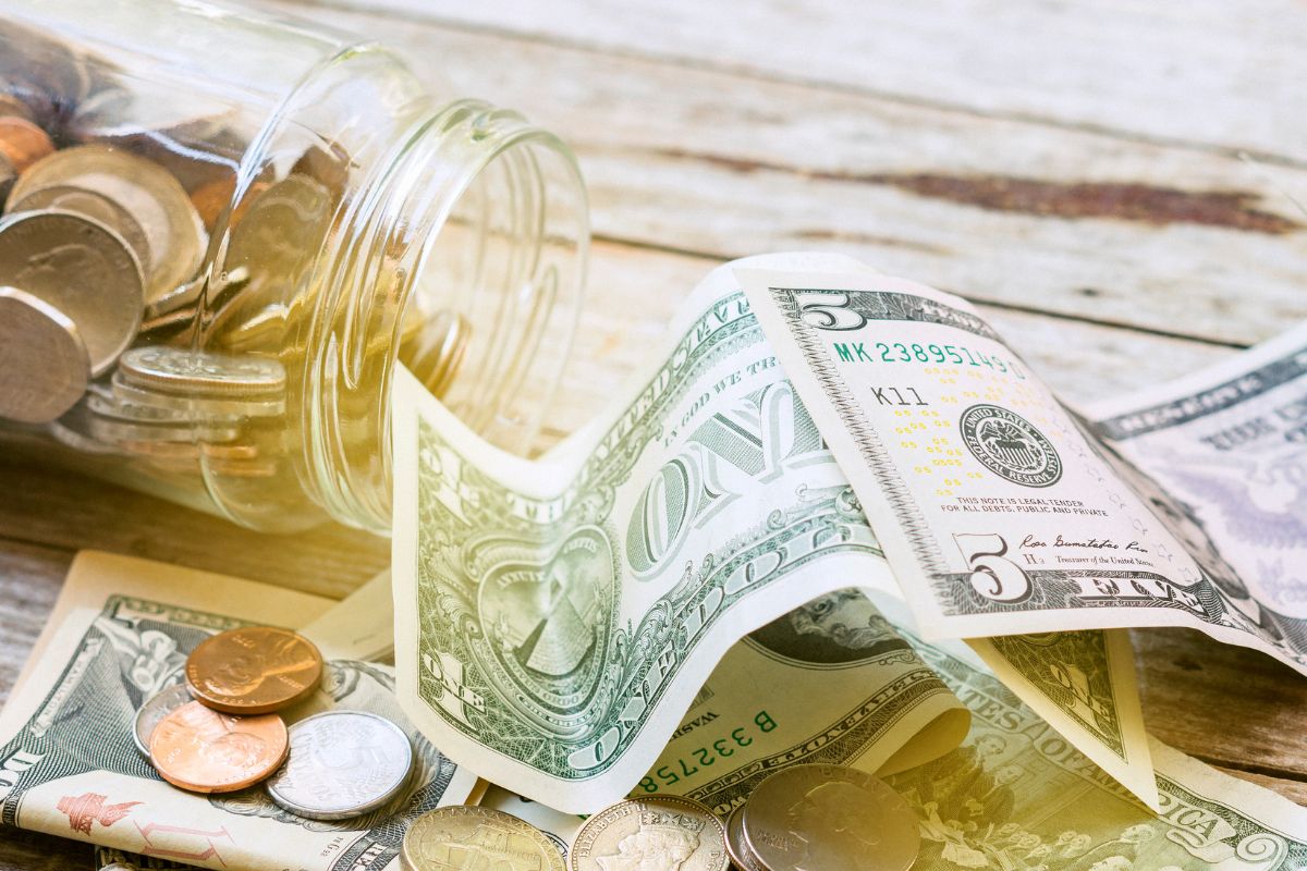 The image shows dollar bills and coins on the wooden table and a jar filled with coins referring to savings not where you want it to be and looking into additional income sources can all help.