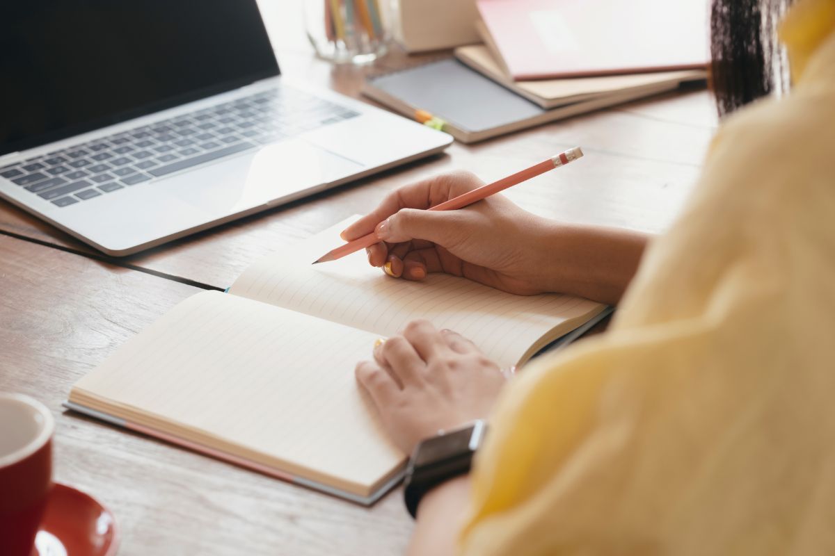 The photo shows a laptop, a mug, and a girl's hand and writing on the notebook referring to learning the steps to financial freedom to control spending and avoid unnecessary debt.