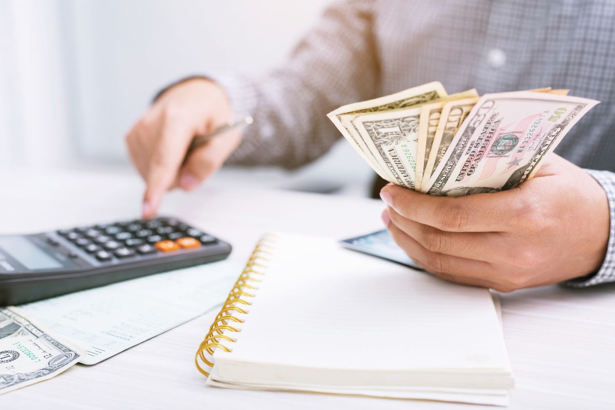 The image tells about a notebook, papers on the table, and a man's hand holding dollar bills while calculating referring to how setting financial goals helps you become more financially secure over time.