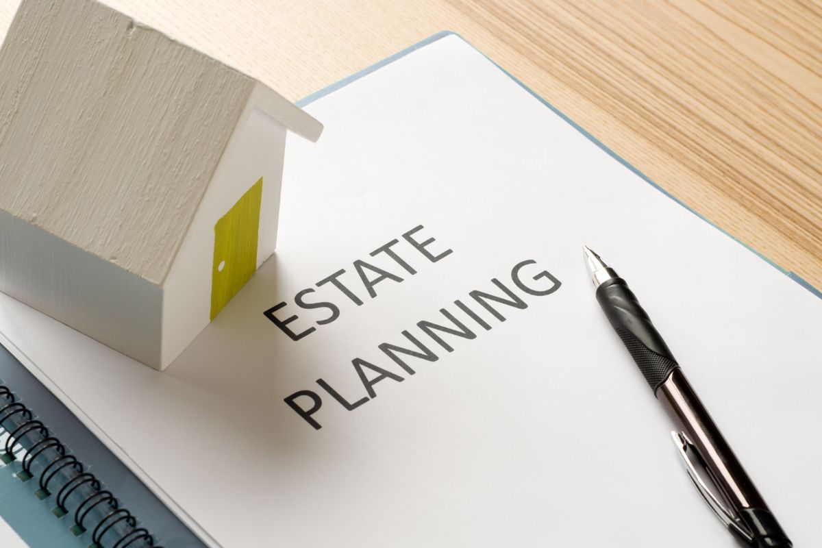 The photo tells about a pen, a wooden house, and a notebook with notes indicating estate planning steps to ensure your money and belongings go where you want after you're gone.