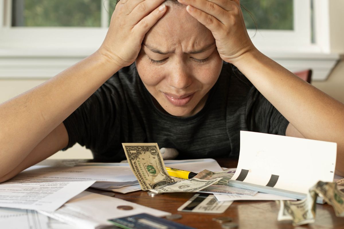 The photo shows papers on the table, dollar bills, credit cards, and a frustrated girl indicating that struggling with money can feel overwhelming, and asking for help from a financial expert can provide guidance.