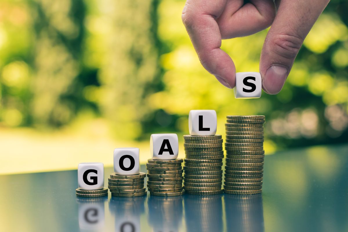 The photo shows coin stocks, wooden cubes with letters, and a man's hand putting last cube indicating to develop a clear vision of financial goals helps you more feel more in control of your future.
